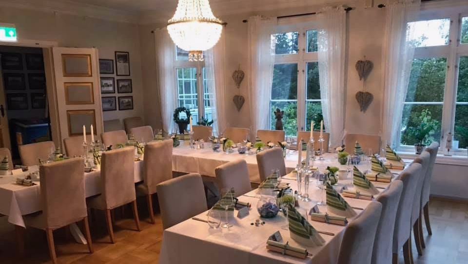 Langbord dekket til selskap i et lyst rom med parkettgulv, lyse stoler og store vinduer med gardiner.